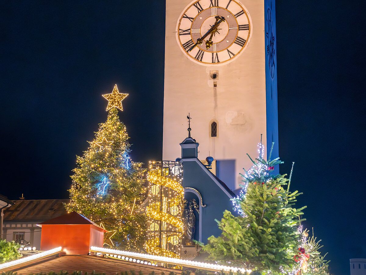 Festlicher Weihnachtsmarkt bei Nacht mit großem, geschmückten Christbaum, leuchtendem Stern und historischem Uhrturm.