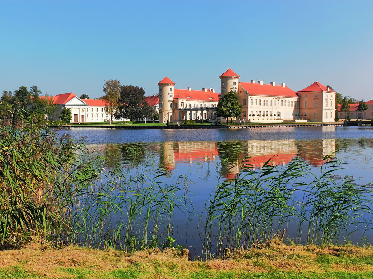 Blick auf das historische Schloss Rheinsberg mit roten Dächern, gespiegelt im blauen See, umgeben von Schilf.