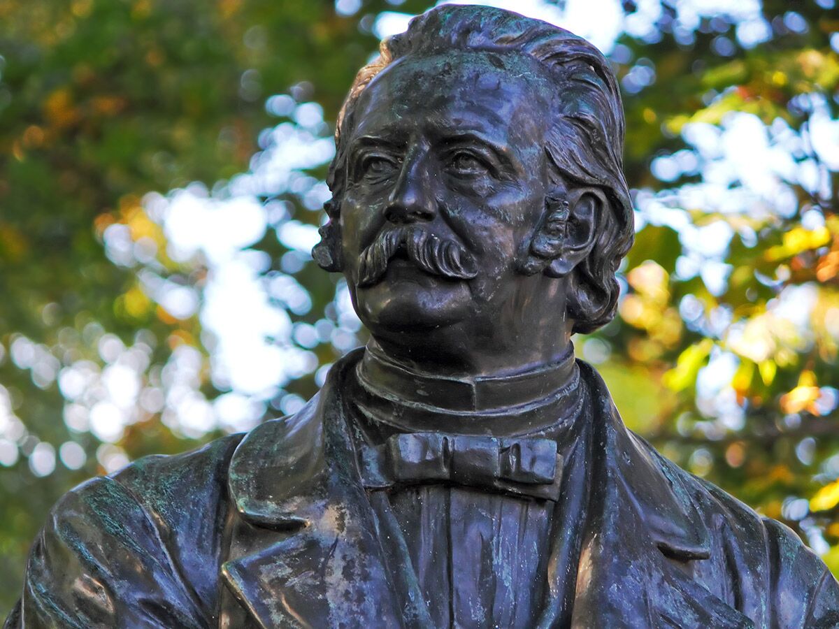 Nahaufnahme einer Bronzestatue eines Mannes mit Schnurrbart und Fliege vor unscharfem grünen Hintergrund.
