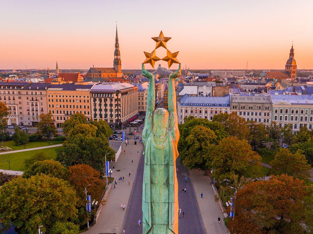 Luftaufnahme des ikonischen Freiheitsdenkmals in Riga bei Sonnenuntergang, goldene Statue, Stadtpanorama mit historischen Gebäuden und Kirchtürmen.