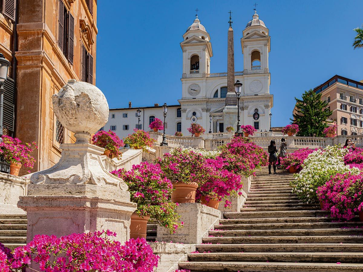 Die Spanische Treppe in Rom ist mit bunten Azaleen geschmückt, im Hintergrund die Kirche Trinità dei Monti unter blauem Himmel.