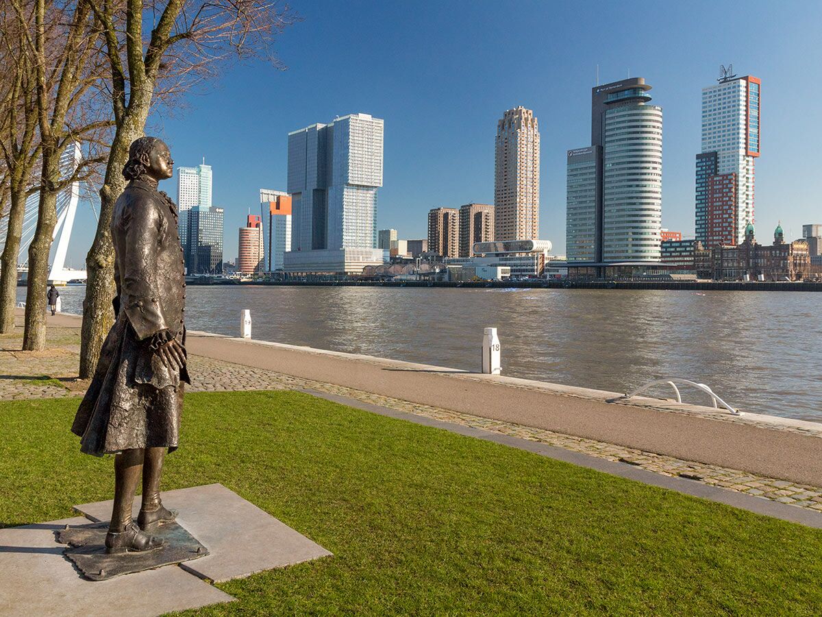 Bronze-Statue von Erasmus blickt auf die Rotterdamer Skyline mit Erasmusbrücke und Nieuwe Maas an einem sonnigen Tag.