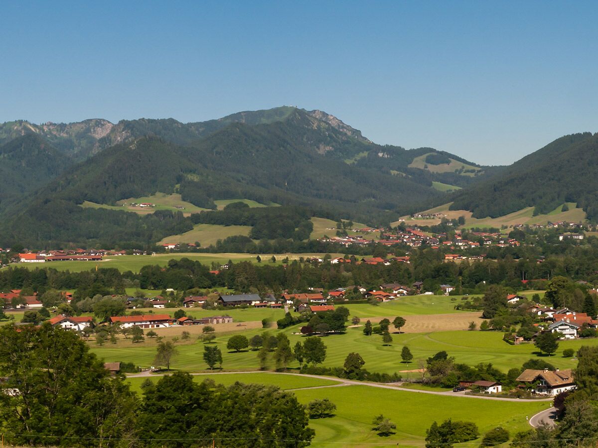 Panoramablick auf ein malerisches Alpendorf mit Häusern, grünen Feldern und bewaldeten Bergen unter blauem Himmel.