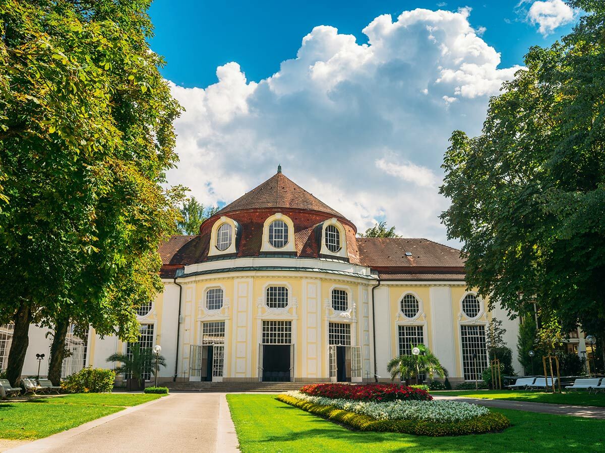 Elegantes Kurhaus mit roter Kuppel im grünen Park. Weiß-gelbes Gebäude, Rundfenster, Rasen, Blumen, Bäume, sonniger Tag.