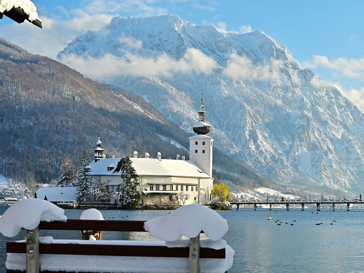 Winterlandschaft mit schneebedecktem Seeschloss Ort am Traunsee und majestätischen Bergen unter blauem Himmel.