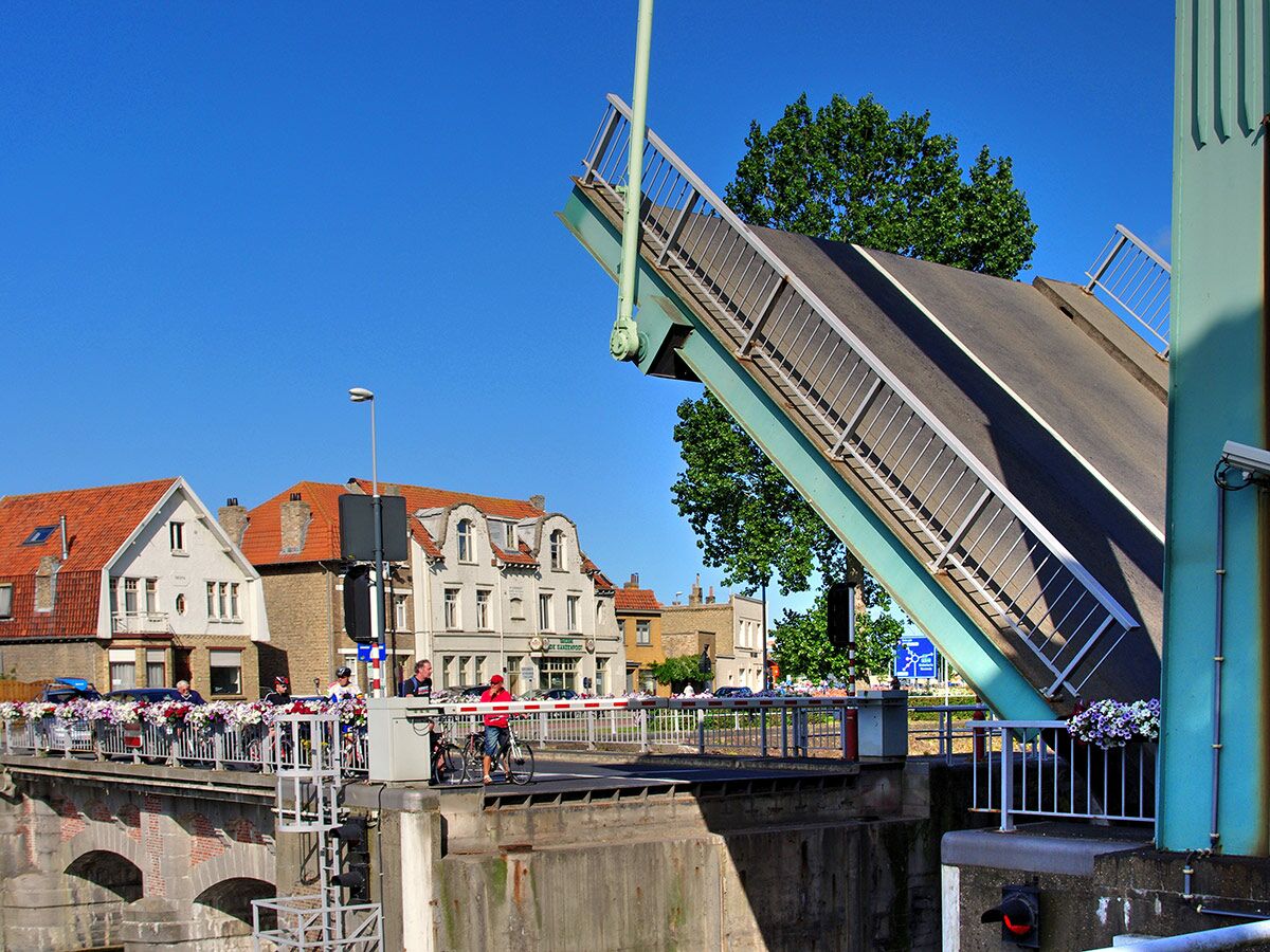 Offene Zugbrücke in einer historischen Stadt mit wartenden Radfahrern, blauer Himmel und roten Dächern.