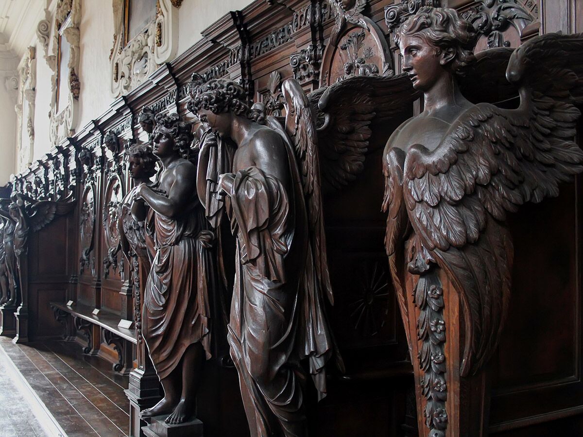 Eine Reihe detailreicher barocker Holzskulpturen von Engeln und mythologischen Figuren in dunklen Chorstühlen einer Kirche.