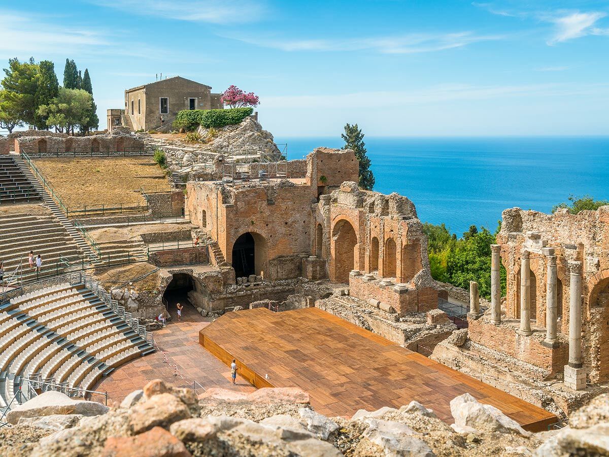 Panorama des antiken Theaters von Taormina mit Ruinen, Sitzreihen, moderner Bühne und dem Ionischen Meer im Hintergrund unter blauem Himmel.