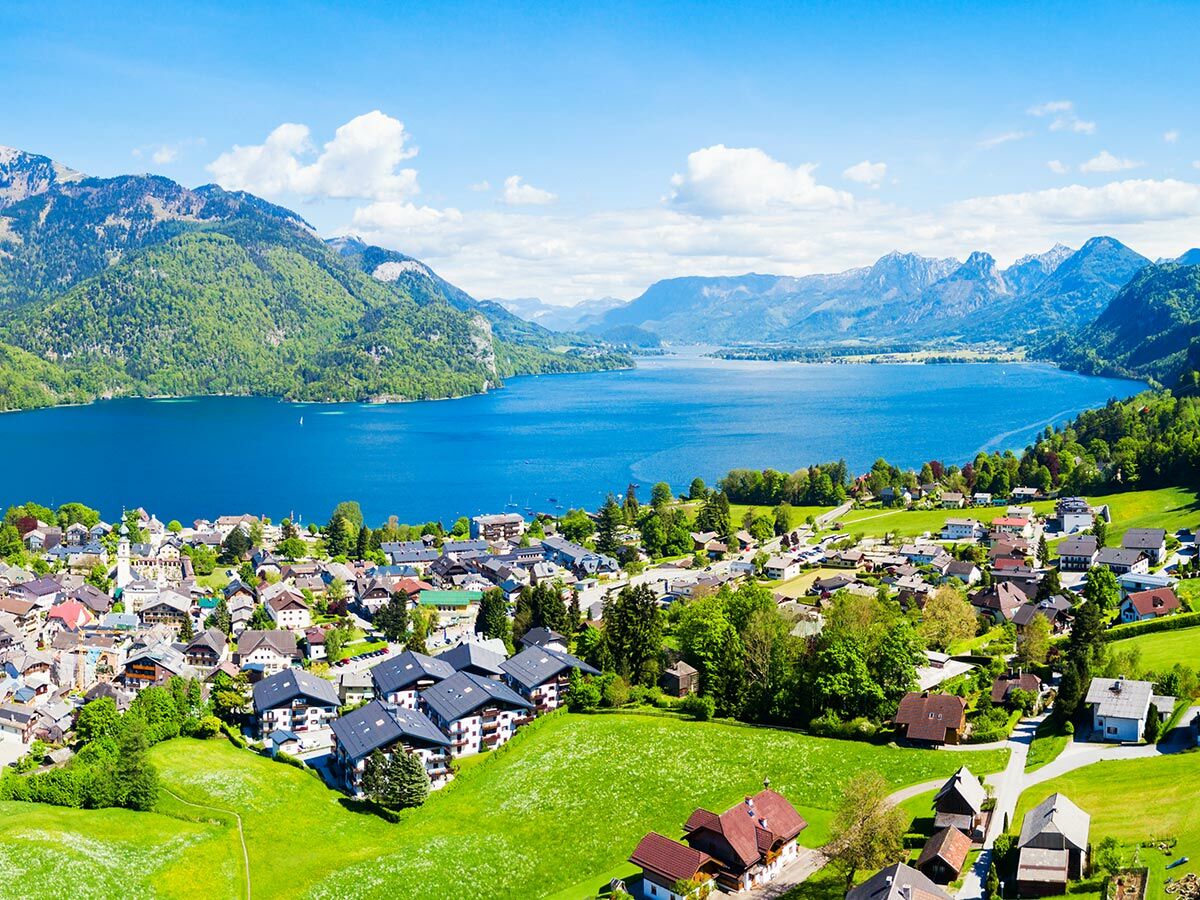 Luftaufnahme eines malerischen Bergdorfs mit Häusern am Ufer eines tiefblauen Sees, umgeben von grünen Bergen unter blauem Himmel.
