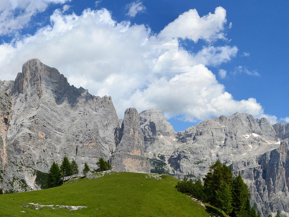 Majestätische Dolomiten mit steilen Felswänden, einem grünen Berghang und vereinzelten Bäumen unter blauem Himmel mit Wolken.