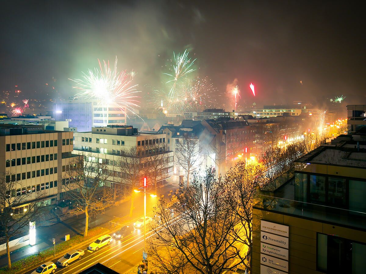 Spektakuläres Feuerwerk über einer nächtlichen Stadt mit beleuchteten Gebäuden und langen Lichtspuren von Autos.