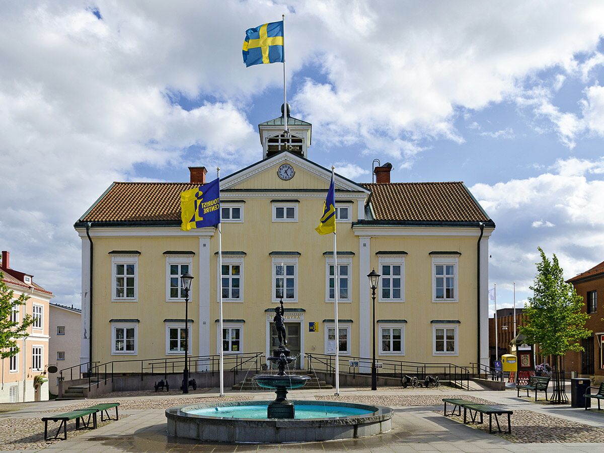 Södra Rådhuset in Lund, Schweden, gelbes historisches Gebäude mit schwedischer Flagge, Uhr und Brunnen auf dem Stadtplatz.