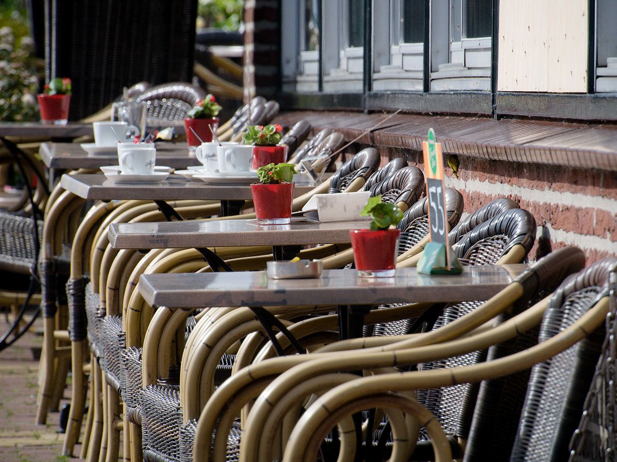 Reihe von leeren Tischen und Stühlen auf einer Straßencafé-Terrasse mit Kaffeetassen und roten Blumentöpfen vor einer Backsteinwand.