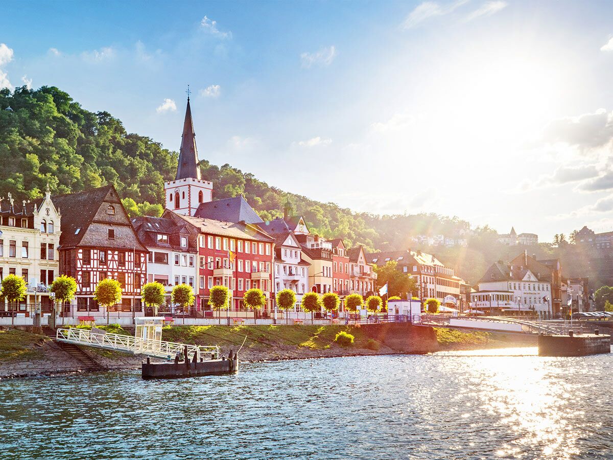 Malerisches Dorf am Rhein mit bunten Fachwerkhäusern, prominenter Kirche und Fähranleger bei Sonnenschein.