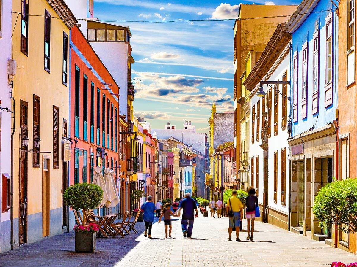 Menschen schlendern durch eine farbenfrohe Altstadtstraße mit malerischen Gebäuden unter blauem Himmel.