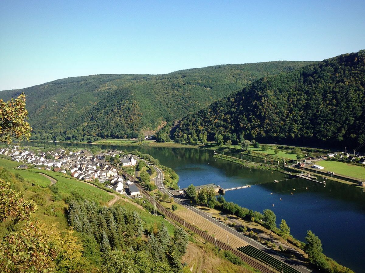 Panoramablick auf ein Flusstal mit malerischem Dorf, grünen Weinbergen, bewaldeten Hügeln und einer Schleuse im Fluss.