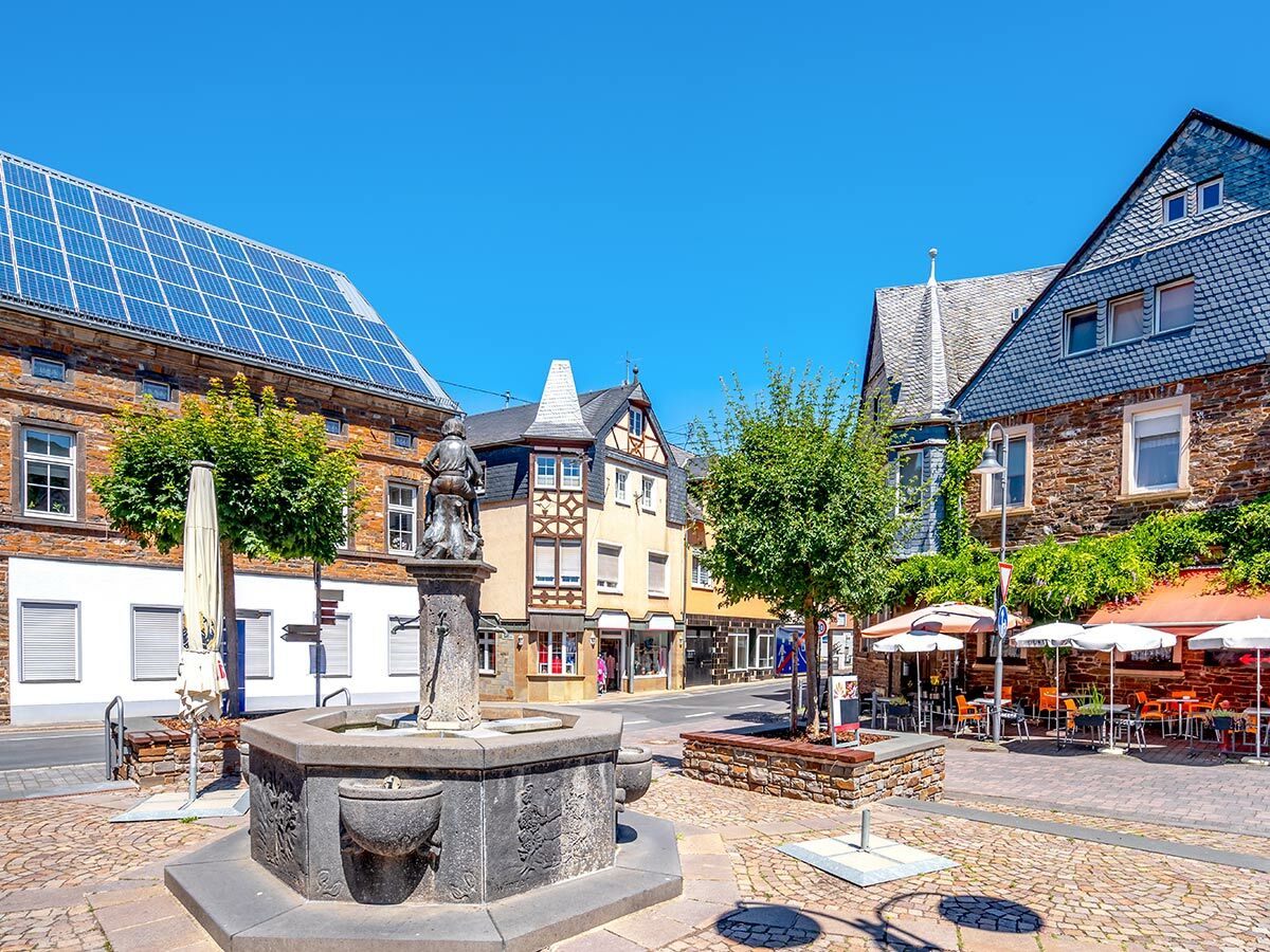 Blick auf einen historischen Stadtplatz mit detailreichem Brunnen, alten Gebäuden, Solarpaneelen und Außengastronomie unter blauem Himmel.