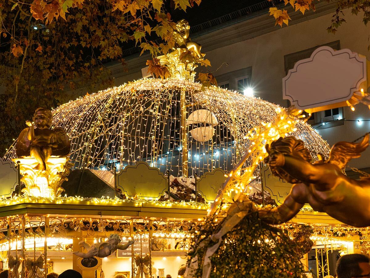 Festlich beleuchteter Weihnachtsmarkt bei Nacht mit goldenen Engeln, Lichterkuppel und Herbstlaub an Bäumen.