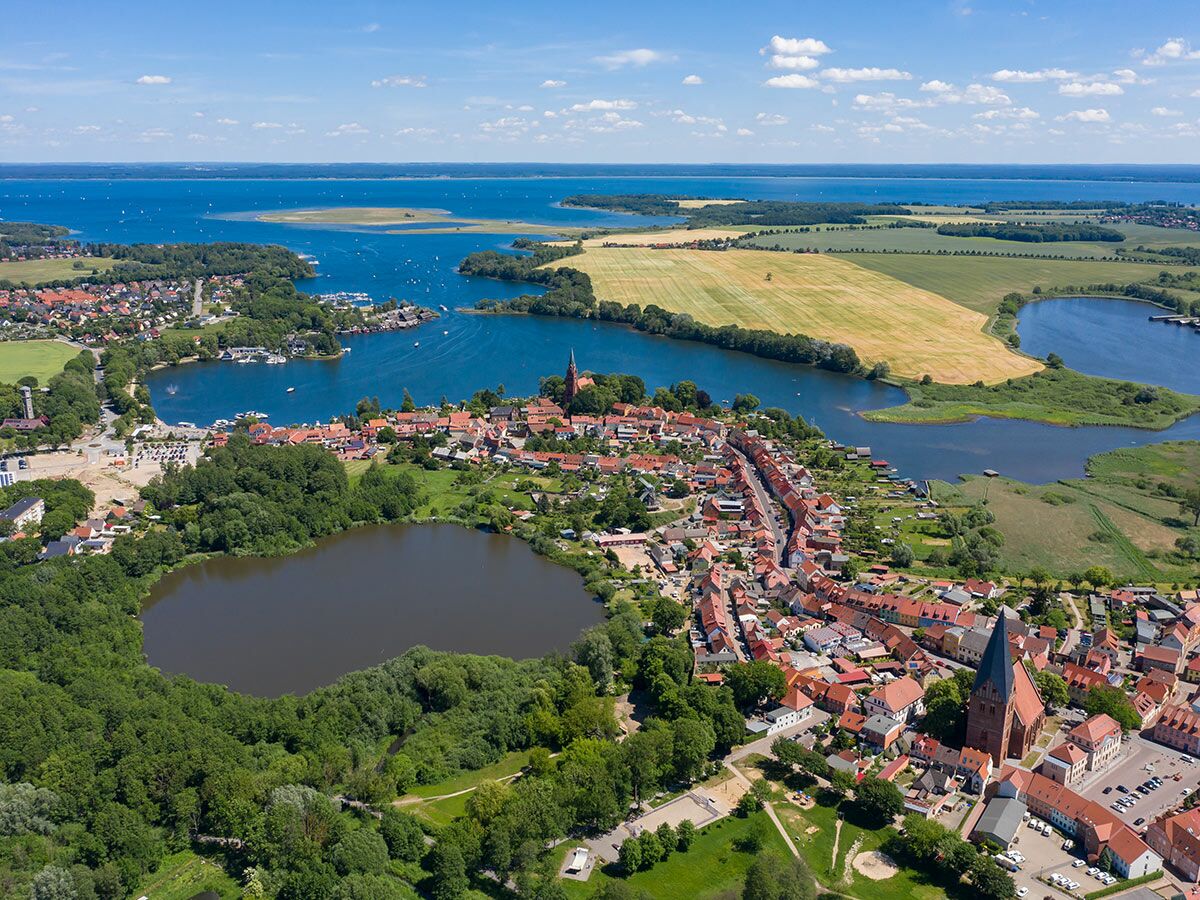 Luftbild einer charmanten Küstenstadt mit roten Dächern, einer Kirche, umgeben von Seen, Feldern und Wäldern unter blauem Himmel.