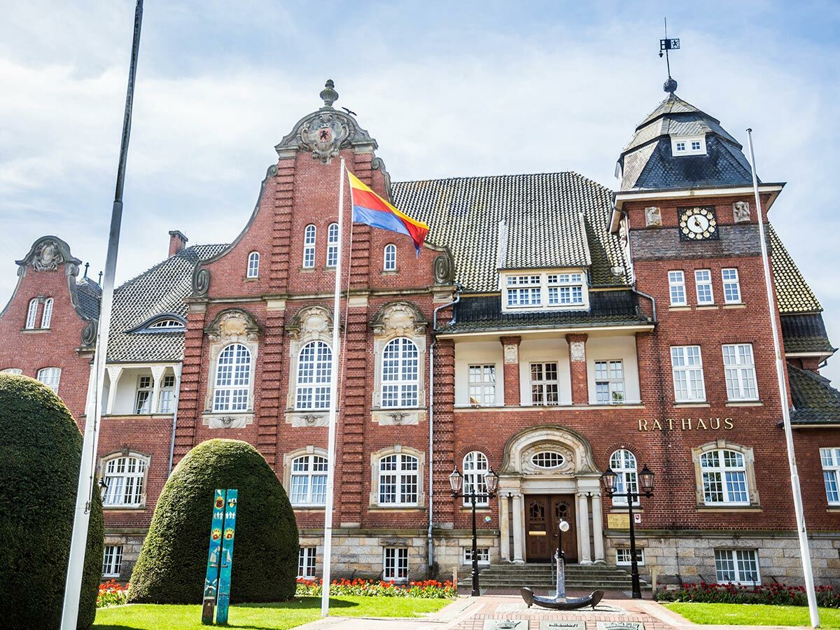 Historisches rotes Backstein-Rathaus mit Turm, Uhr, wehender Flagge, Anker-Skulptur. Vorplatz mit Büschen und Blumen.