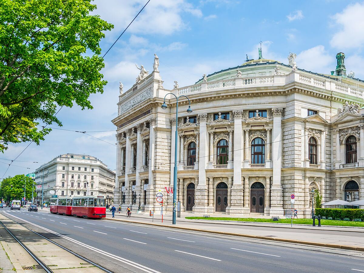 Blick auf die Wiener Staatsoper an sonnigem Tag mit roter Straßenbahn und Passanten auf der Ringstraße in Wien.