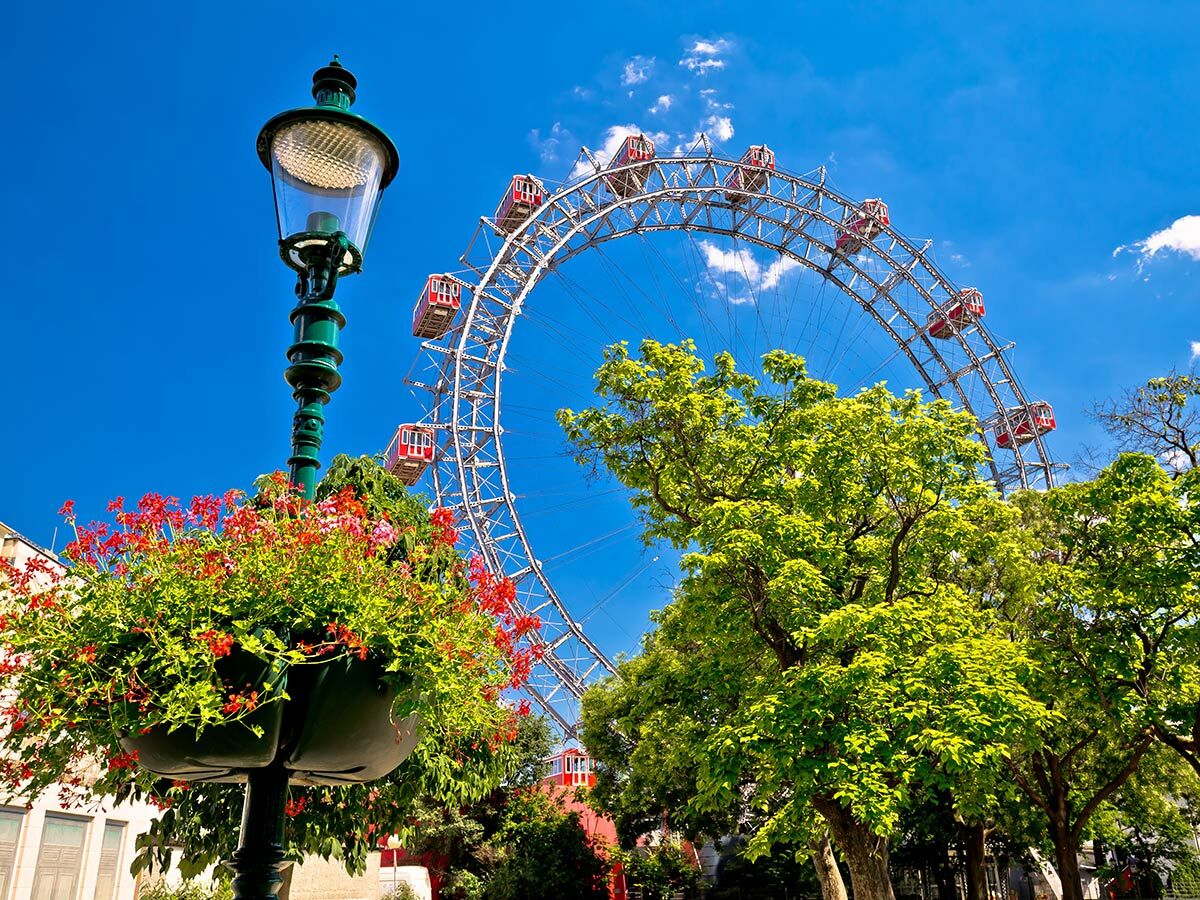 Grüne Laterne mit roten Blumen vor dem Wiener Riesenrad und Bäumen unter blauem Himmel im Prater.