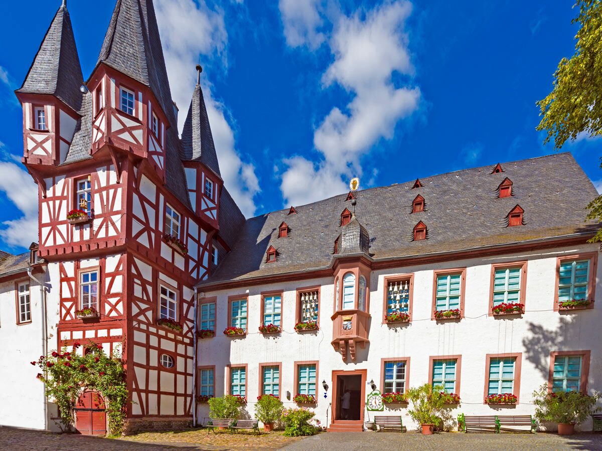 Blick auf ein historisches deutsches Fachwerkhaus mit roten Balken, weißen Wänden, Türmen und Blumenkästen unter blauem Himmel.