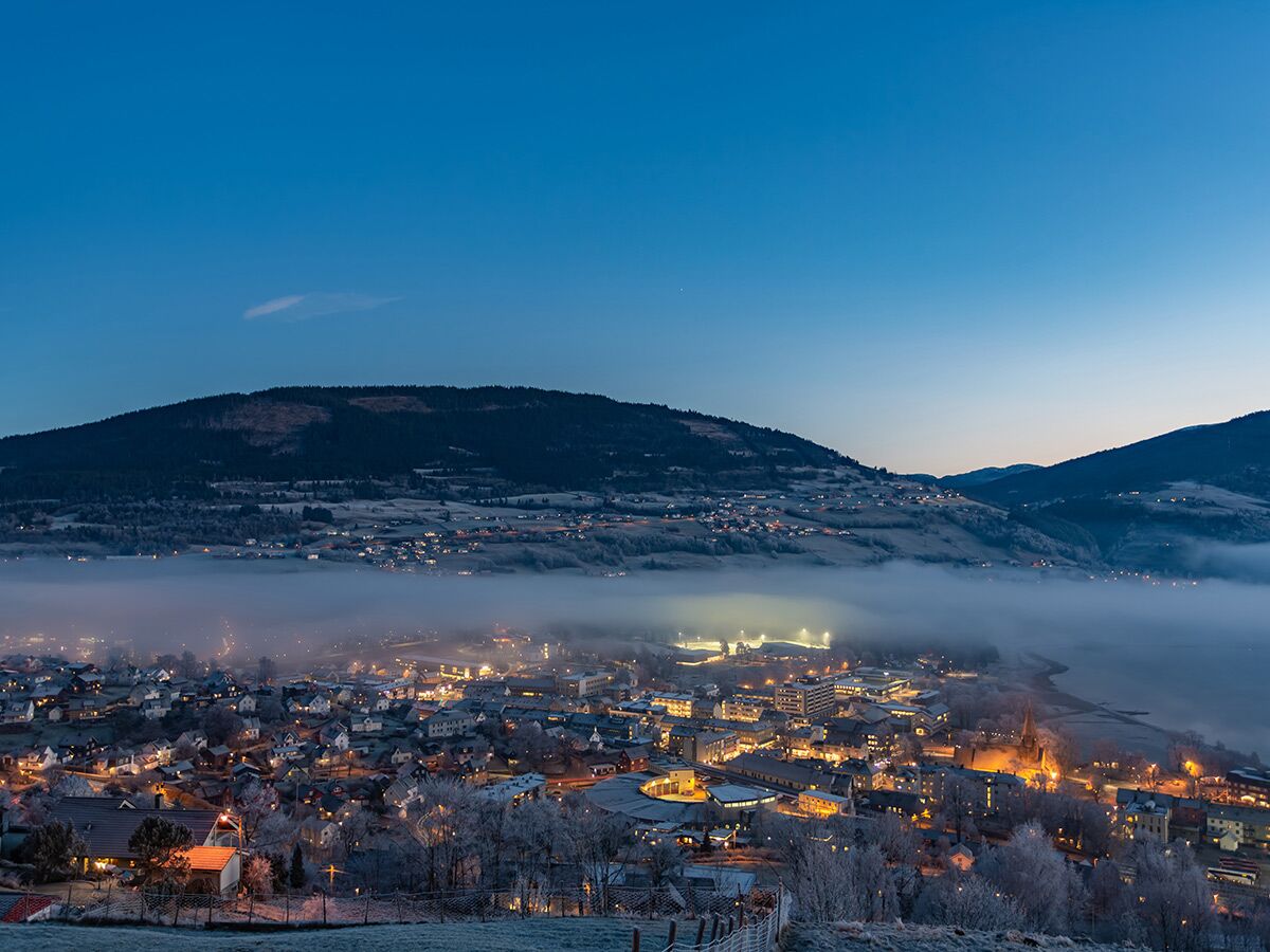 Panoramablick auf eine winterliche Stadt im Tal, eingehüllt in Nebel, mit beleuchteten Häusern und Bergen bei Dämmerung.