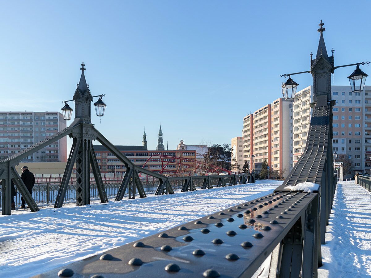 Verschneite Eisenbrücke mit Laternen an einem sonnigen Wintertag in der Stadt, im Hintergrund hohe Gebäude und Kirchtürme.