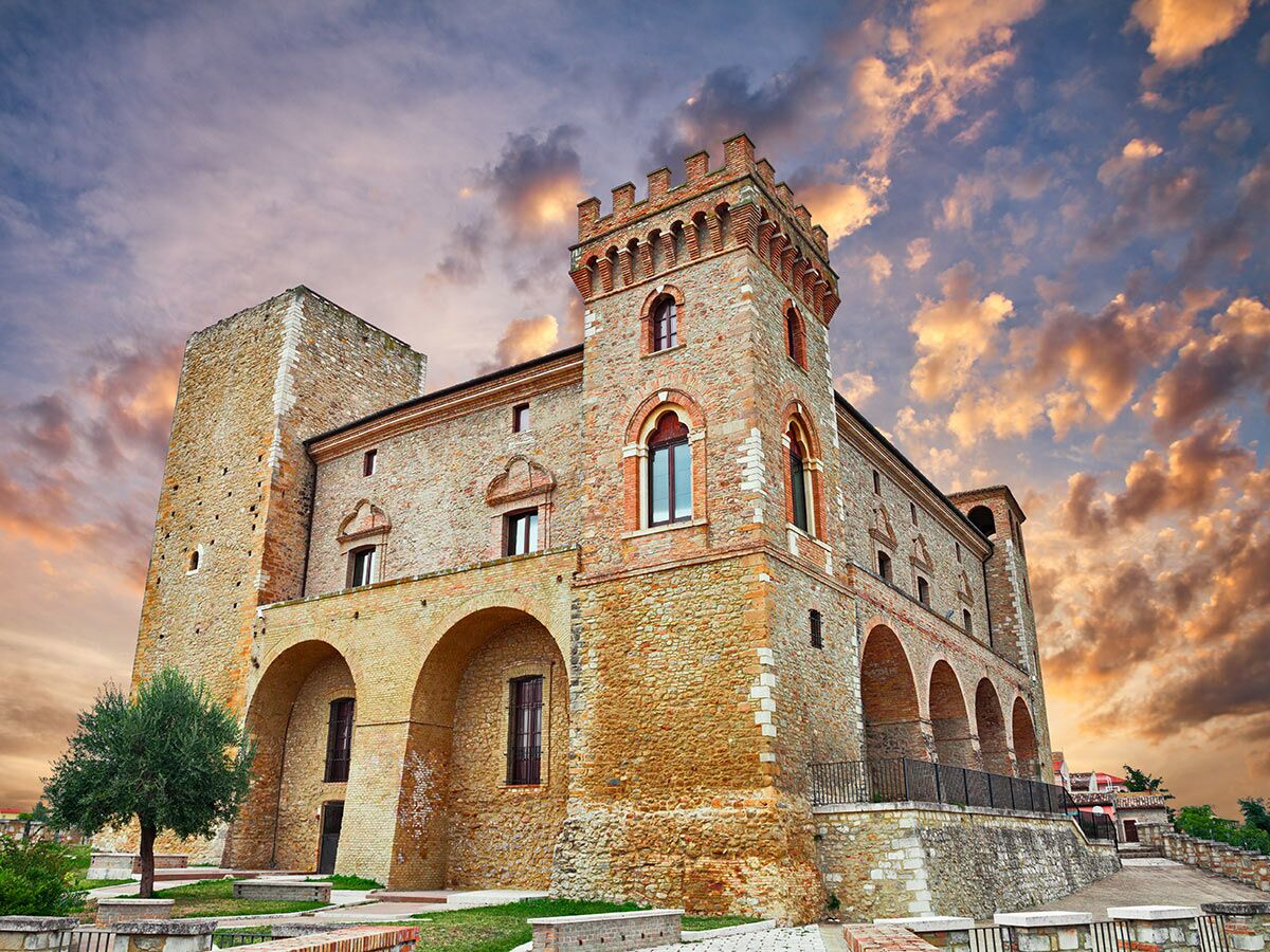 Blick auf ein altes italienisches Schloss mit hohen Türmen und Zinnen, aus Stein und Ziegeln, unter einem dramatischen Wolkenhimmel bei Sonnenuntergang.