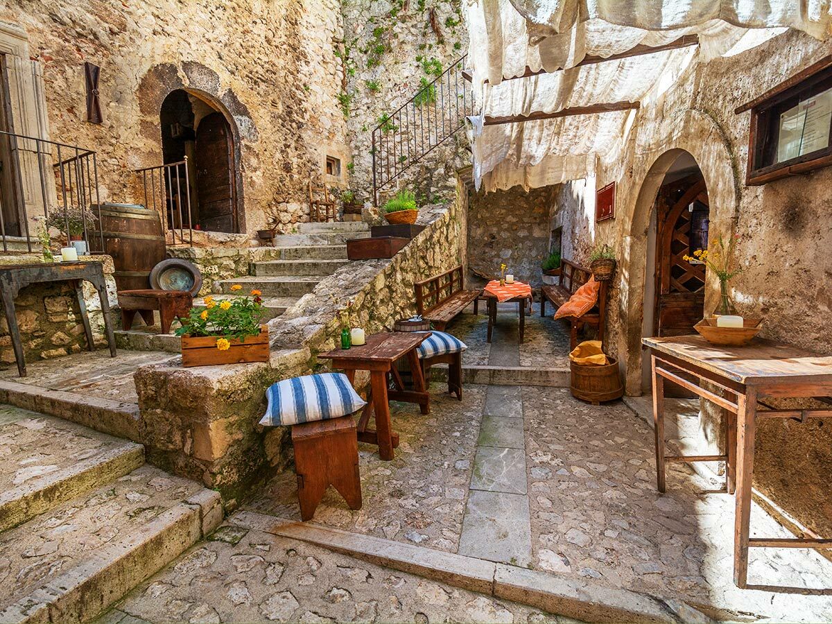 Ein charmanter mediterraner Innenhof mit alten Steinwänden, Holztischen, Bänken und Stufen, geschmückt mit Pflanzen und Textilien im Sonnenlicht.