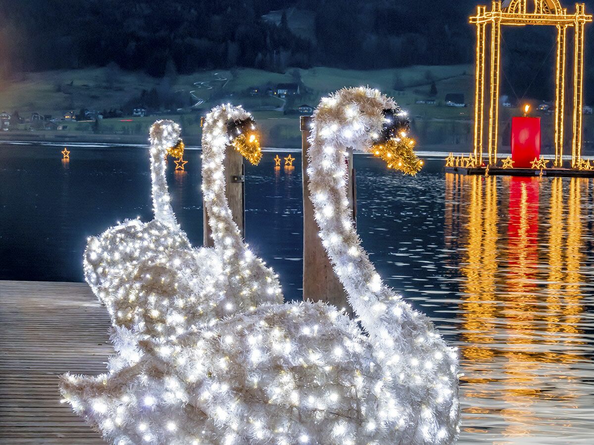 Beleuchtete Schwan-Dekorationen auf einem Steg am See, mit einem roten Kerzen-Lichterbogen und schwebenden Sternen im Wasser bei Nacht.