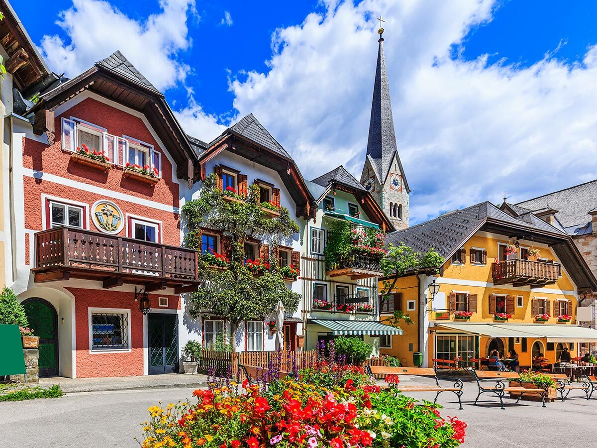 Buntes Hallstatt Dorf mit traditionellen Häusern, Blumen, Kirchturm und Alpenblick unter blauem Himmel in Österreich.