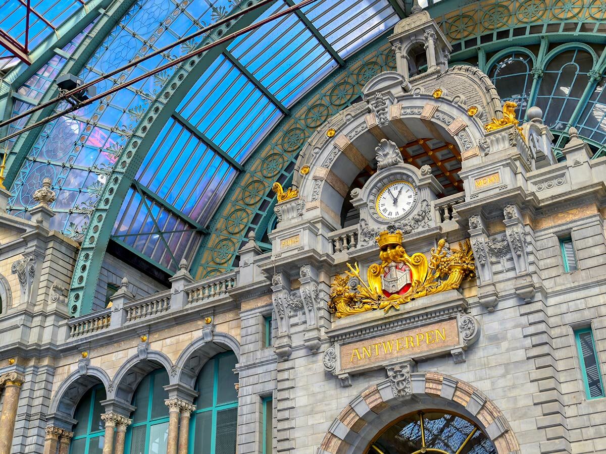 Opulente Architektur des Hauptbahnhofs Antwerpen mit Uhr, goldener Wappenverzierung und blauer Glaskuppel.