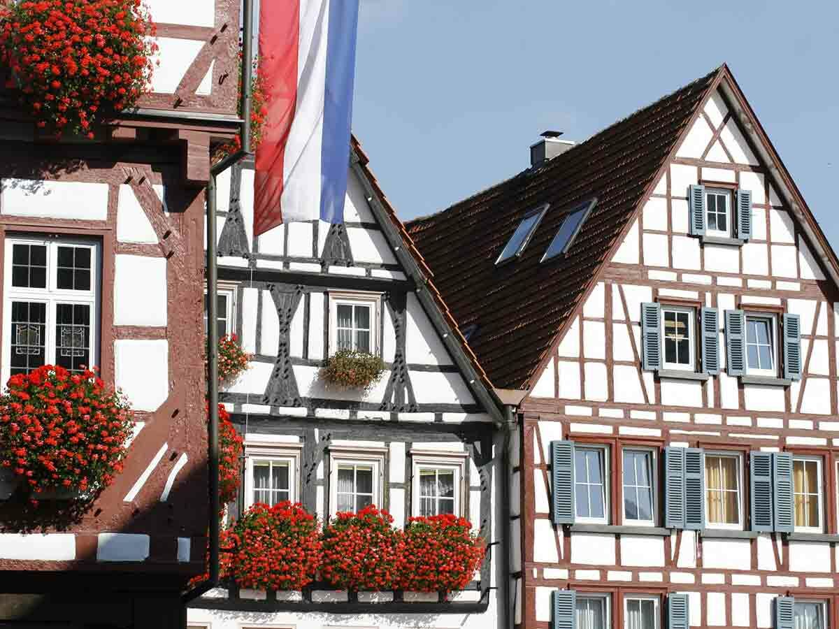 Detailansicht traditioneller Fachwerkhäuser mit roten Geranien in Blumenkästen und einer rot-weiß-blauen Flagge unter blauem Himmel.