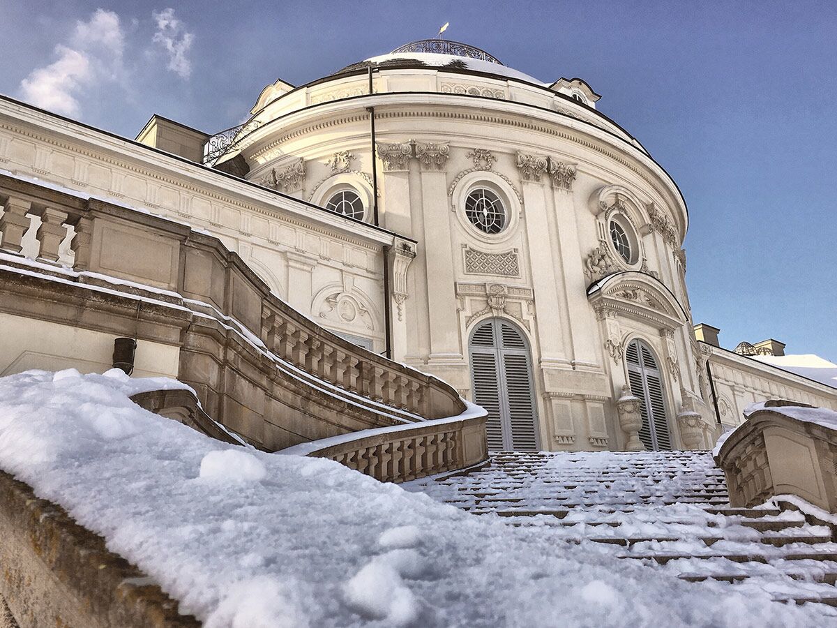 Weiße Fassade des Stupinigi Palastes in winterlicher Schneelandschaft mit imposanter Treppe unter blauem Himmel.