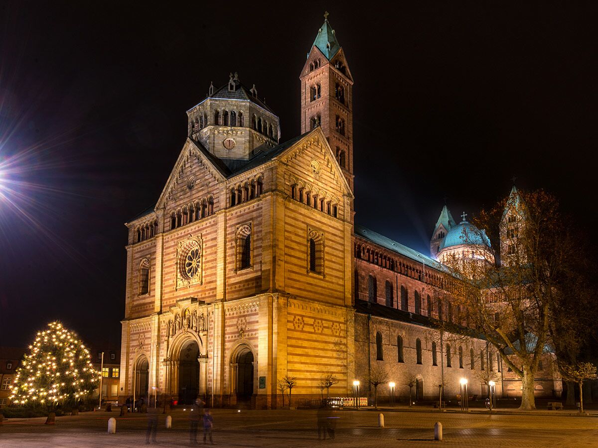 Der Speyerer Dom ist nachts warm beleuchtet, mit einem festlich geschmückten Weihnachtsbaum auf dem Vorplatz.