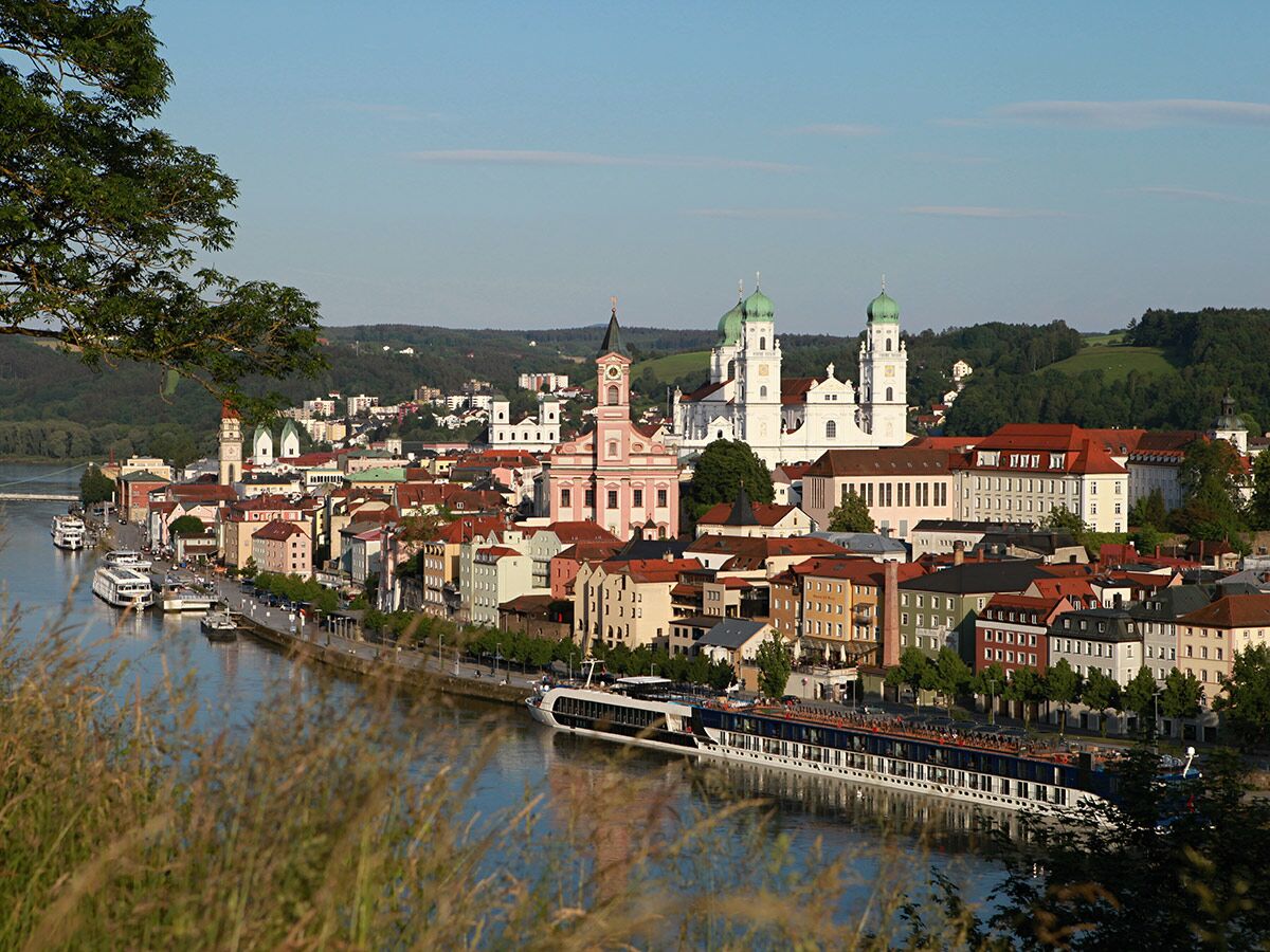 Panorama von Passau mit Dom St. Stephan, pinker Kirche, bunten Häusern und Flusskreuzfahrtschiffen an der Donau.