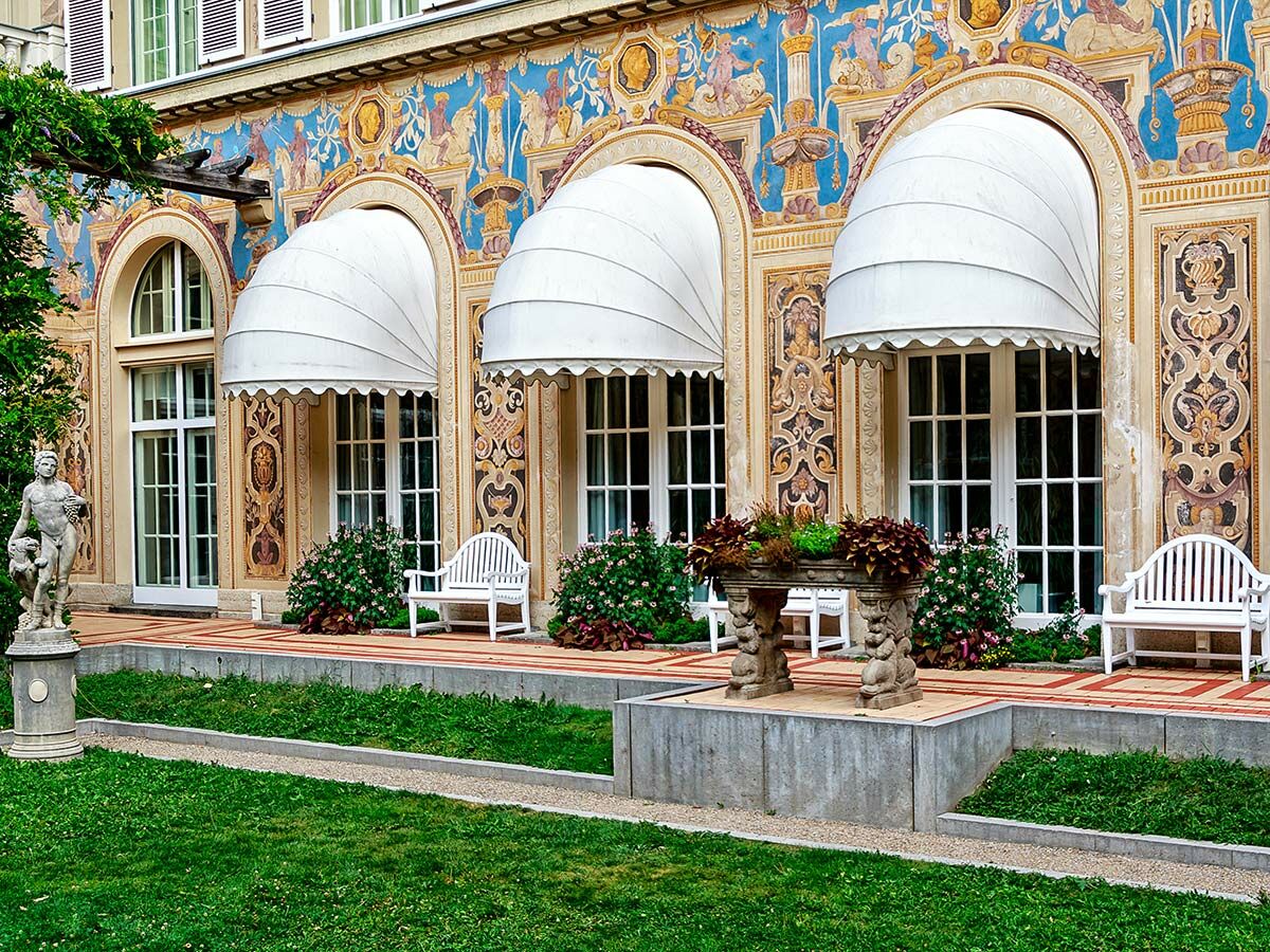 Prächtige Fassade einer historischen Villa mit bemalten Fresken, weißen Markisen, Garten und klassischer Statue.