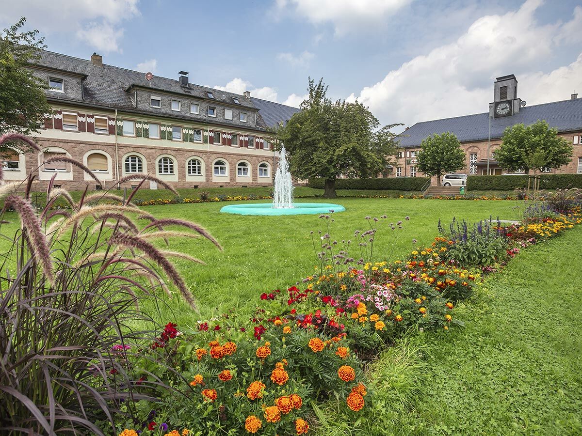 Historisches Gebäude mit Uhr-Turm in einem gepflegten Garten mit farbenfrohen Blumenbeeten und einem Brunnen unter blauem Himmel.