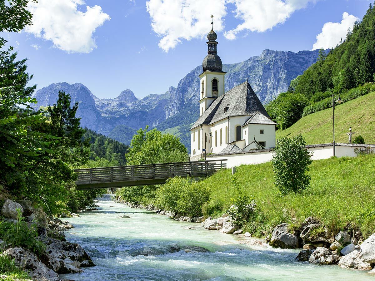 Idyllische St. Sebastian Kirche in Ramsau, Bayern, umgeben von grünen Alpenbergen, einem türkisen Fluss und einer Holzbrücke.