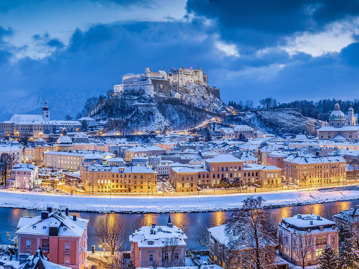 Panoramablick auf Salzburg im Winter bei Dämmerung, verschneite Häuser, Hohensalzburg Festung und Salzach Fluss leuchten.