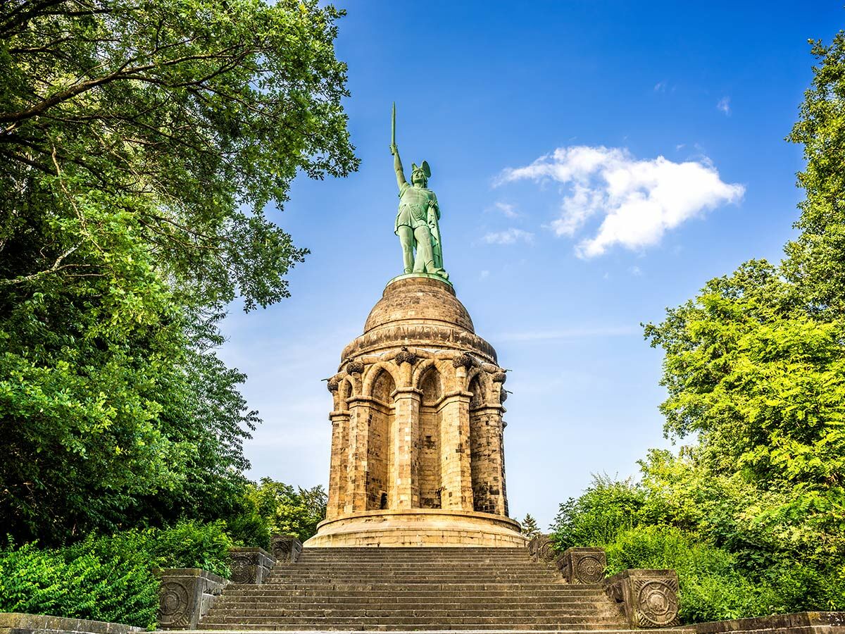 Das Hermannsdenkmal in Detmold zeigt die Statue des Cheruskerfürsten Arminius mit erhobenem Schwert auf einem runden Podest, umgeben von Bäumen.