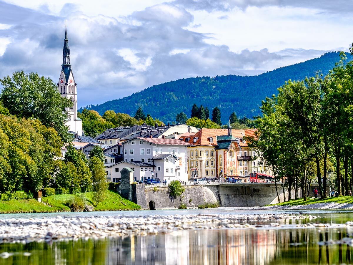 Historische europäische Stadt am Fluss, mit hoher Kirchturmspitze, charmanten Häusern, Brücke, bewaldeten Bergen und Reflexionen.