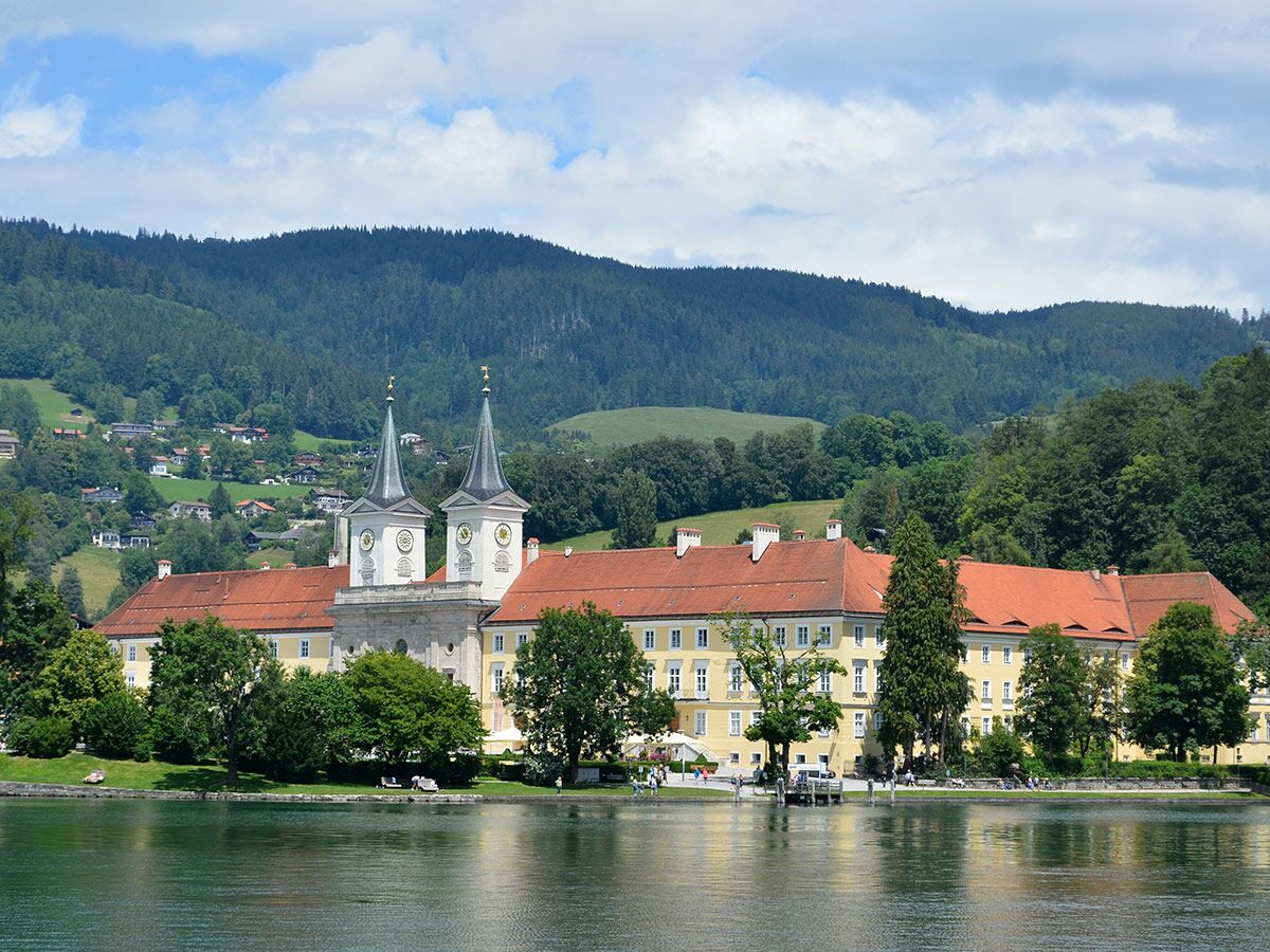 Panoramablick auf ein gelbes Klostergebäude mit roten Dächern und zwei Türmen direkt an einem See, umgeben von grünen Bergen und Wald.