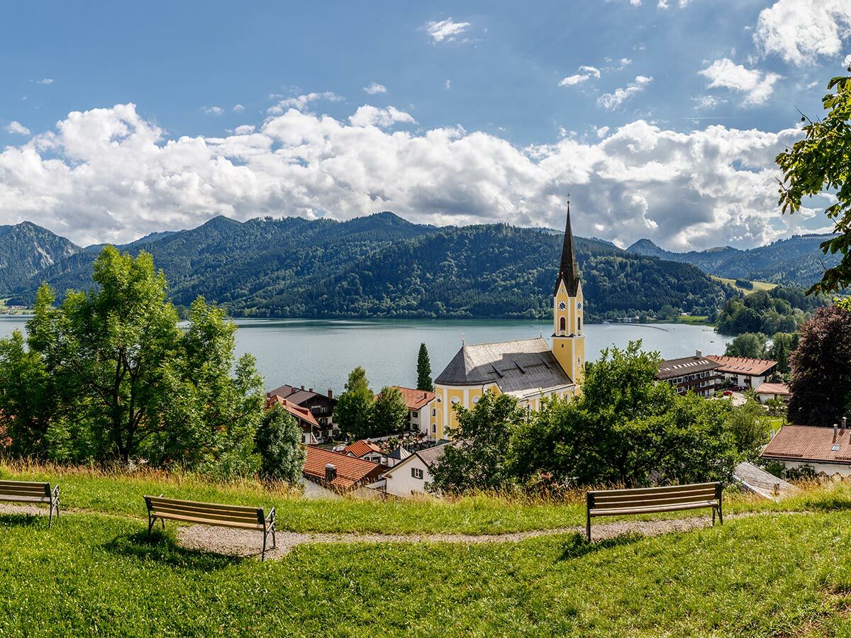 Idyllische Landschaft am Tegernsee mit gelber Dorfkirche, Häusern, See und bewaldeten Bergen unter blauem Himmel. Zwei Parkbänke.