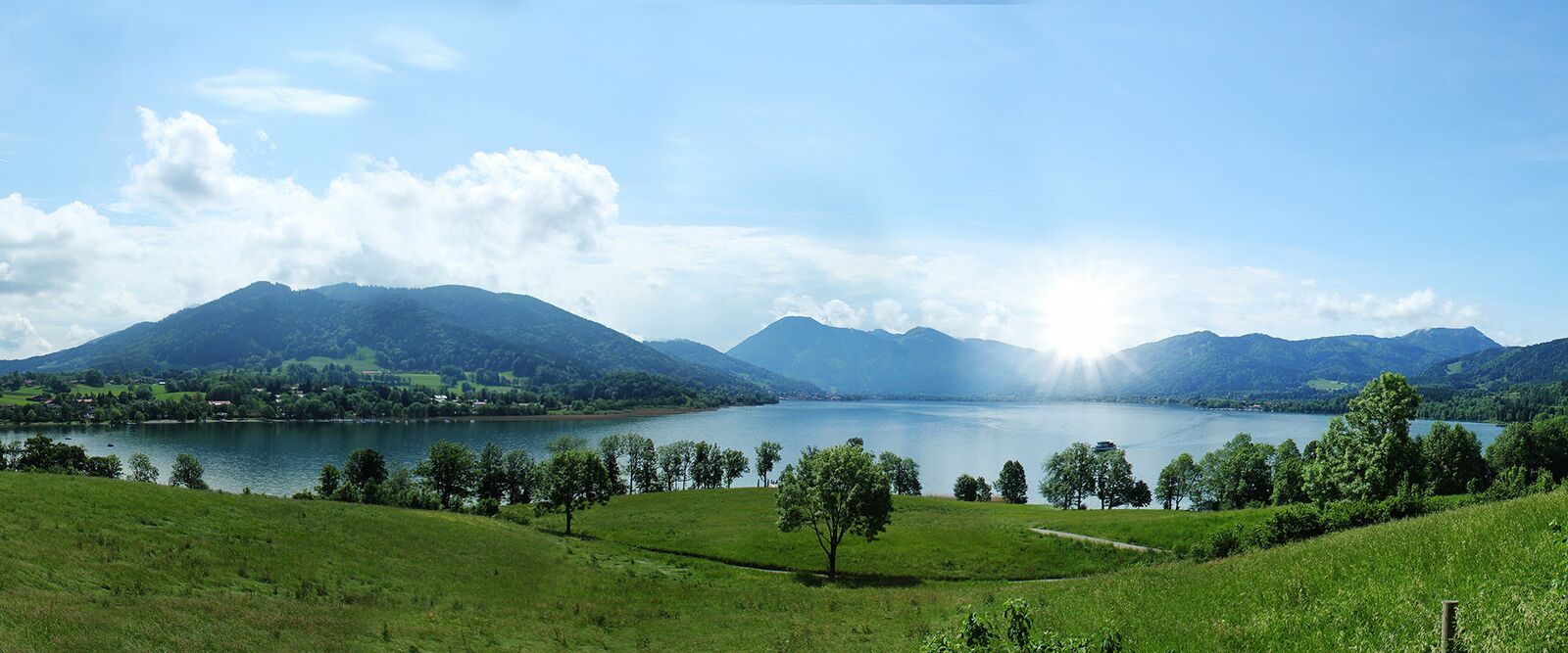 Panorama eines alpinen Sees mit grünen Wiesen, bewaldeten Bergen und hellem Sonnenschein unter blauem Himmel und Wolken.