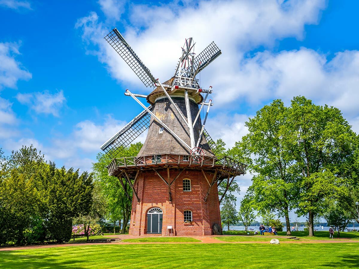 Majestätische Backstein-Windmühle mit Strohdach und Flügeln in einem grünen Park unter blauem Himmel mit Wolken, am Wasser.