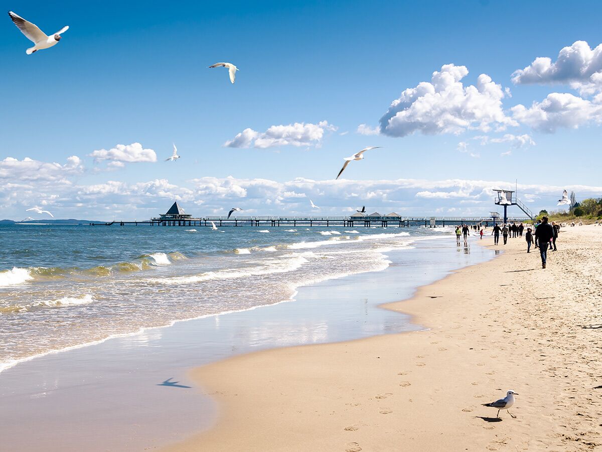 Sonnenverwöhnter Strand mit Menschen, die spazieren gehen, Möwen fliegen, und einer langen Seebrücke mit markantem Gebäude in der Ferne.