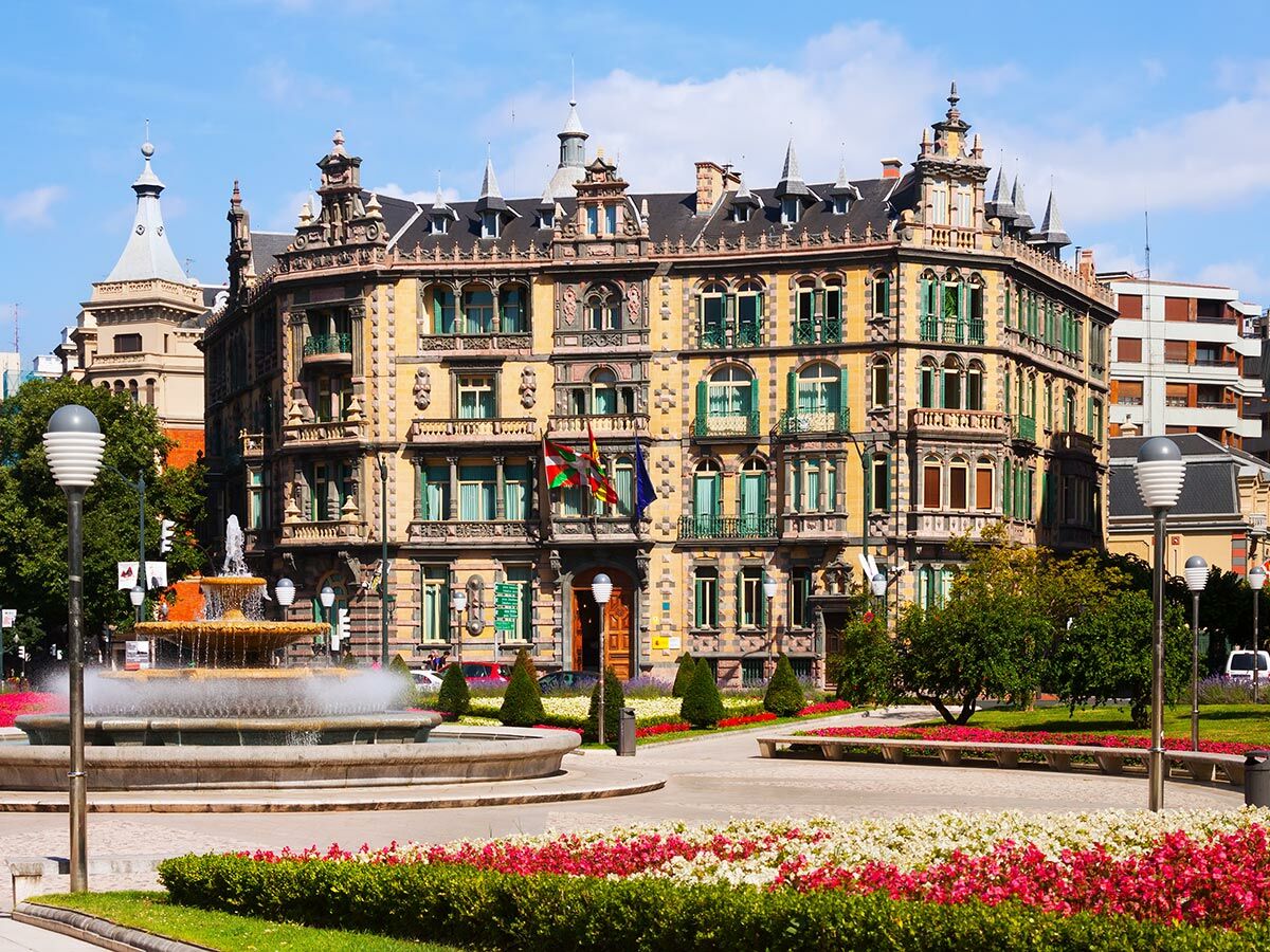Ornates Gebäude des Provinzrates von Bizkaia in Bilbao, Spanien, mit grünen Fensterläden, Brunnen und Blumenbeeten bei Sonne.